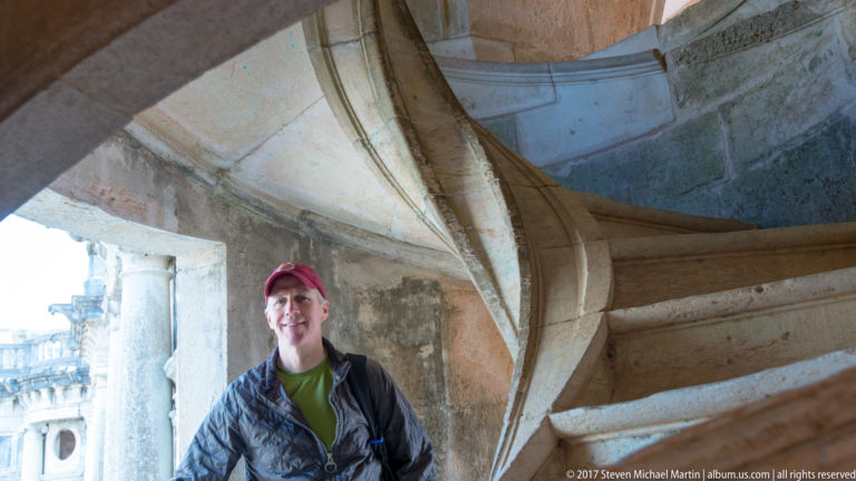 Visitor in a red cap stands beneath a curving stone spiral staircase beside a bright cloister opening.