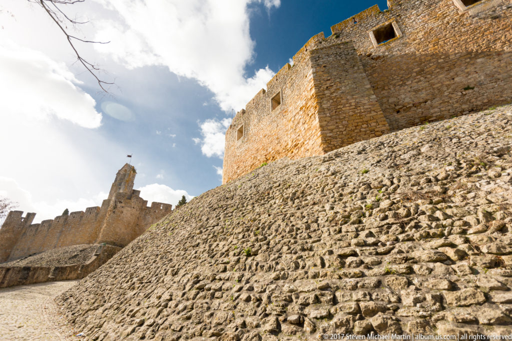 Steep low-angle view up a rough sloped stone defense toward high crenellated castle walls with rectangular openings.