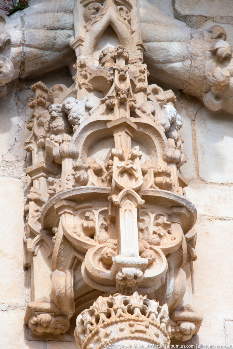Elaborate carved stone crest with scrolls, figures, heraldic forms, and a cross-like emblem above an opening.