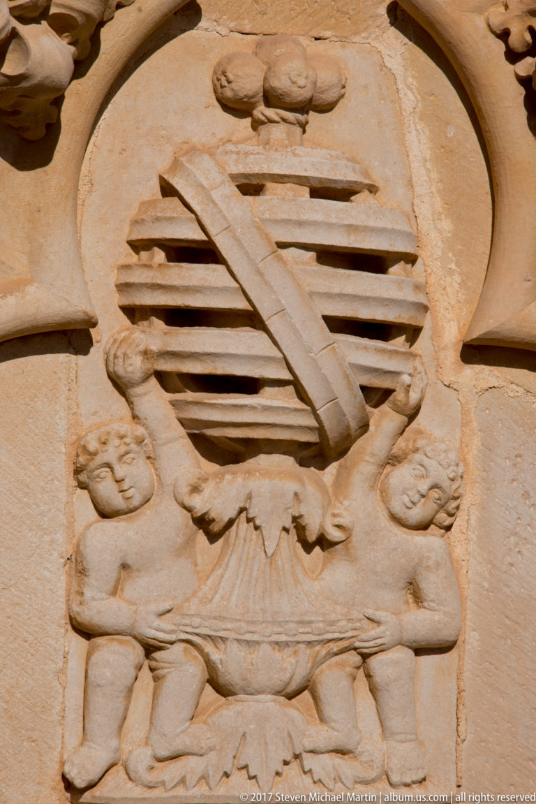 Weathered stone heraldic relief with diagonal bands across a central shield-like field and small figures below.