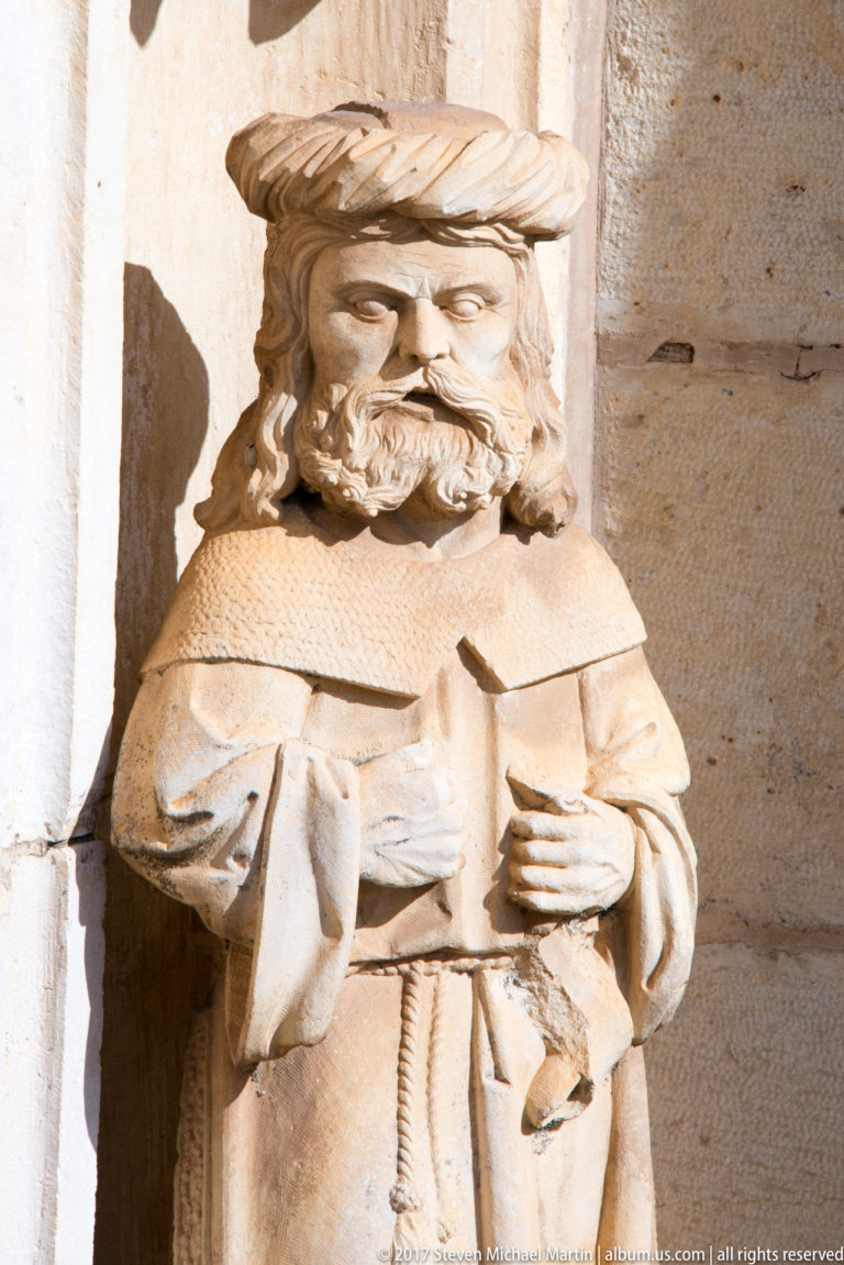 Carved bearded male figure with wrapped headwear and layered clothing, holding a small object against his chest.