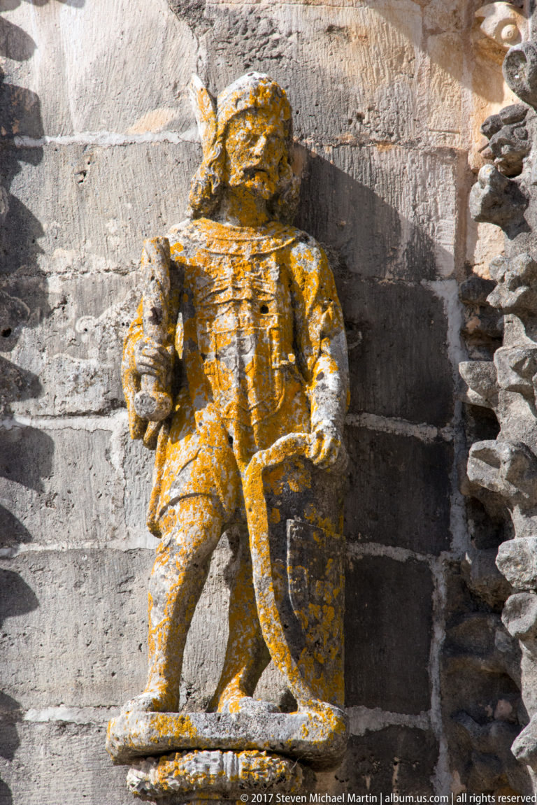 Weathered armored male stone figure standing in a niche, with lichen across the head, torso, and legs.