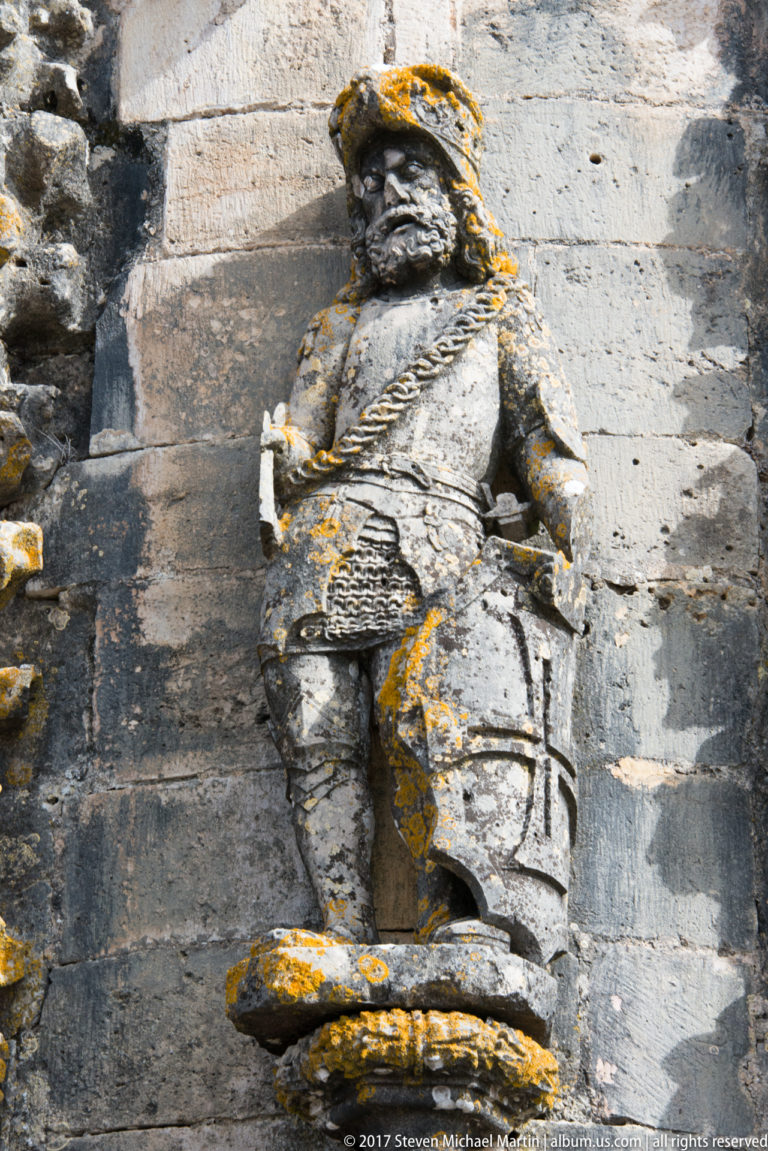 Weathered bearded male stone figure in historical dress standing in a shallow exterior niche.