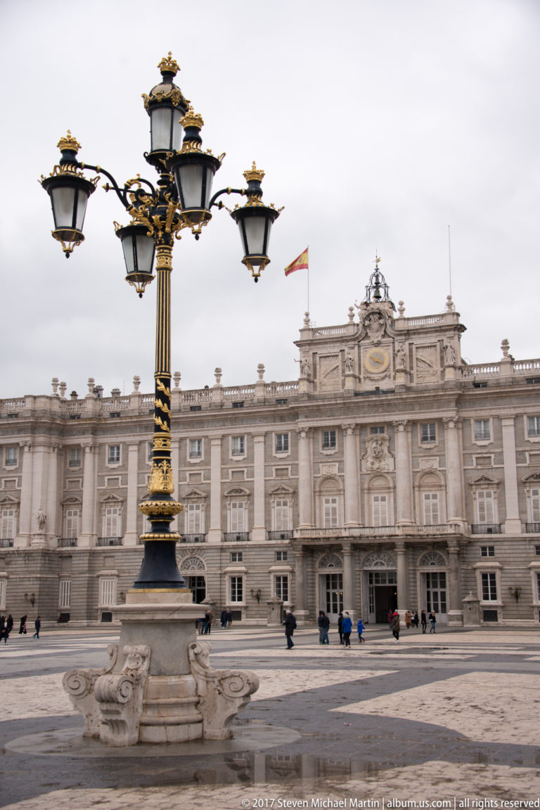 Royal Palace of Madrid across a broad paved square, with ornate black-and-gold street lamp prominent in the foreground.