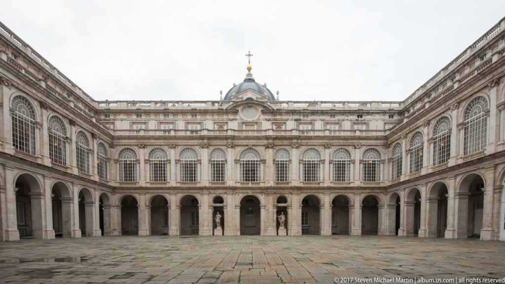 Symmetrical stone courtyard of Madrid’s Royal Palace with arcades, tall windows, central pavilion, and empty paving.
