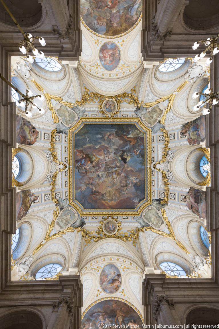 Ornate palace ceiling with central mythological fresco, gilded stucco, oval medallions, and white sculpted figures.