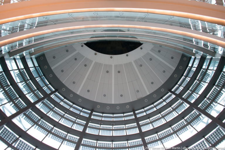 Upward view inside a circular modern atrium with stacked balconies, glass, radial ceiling panels, and a curved foreground rail.