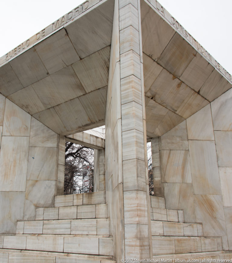 Interior of an open pale-marble pavilion with thick rectangular piers, broad roof slabs, steps, and trees visible through openings.
