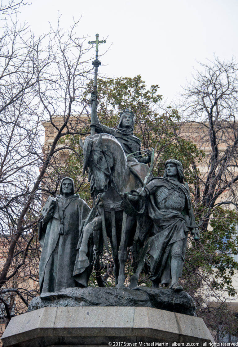 Bronze equestrian monument with a mounted man at center and several accompanying figures, seen against leafless trees.