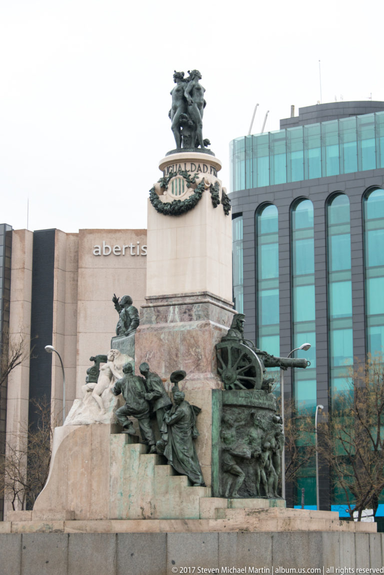 Large civic monument with bronze figure groups, cannon-like forms, and a pedestal inscribed “IGUALDAD,” backed by modern buildings.