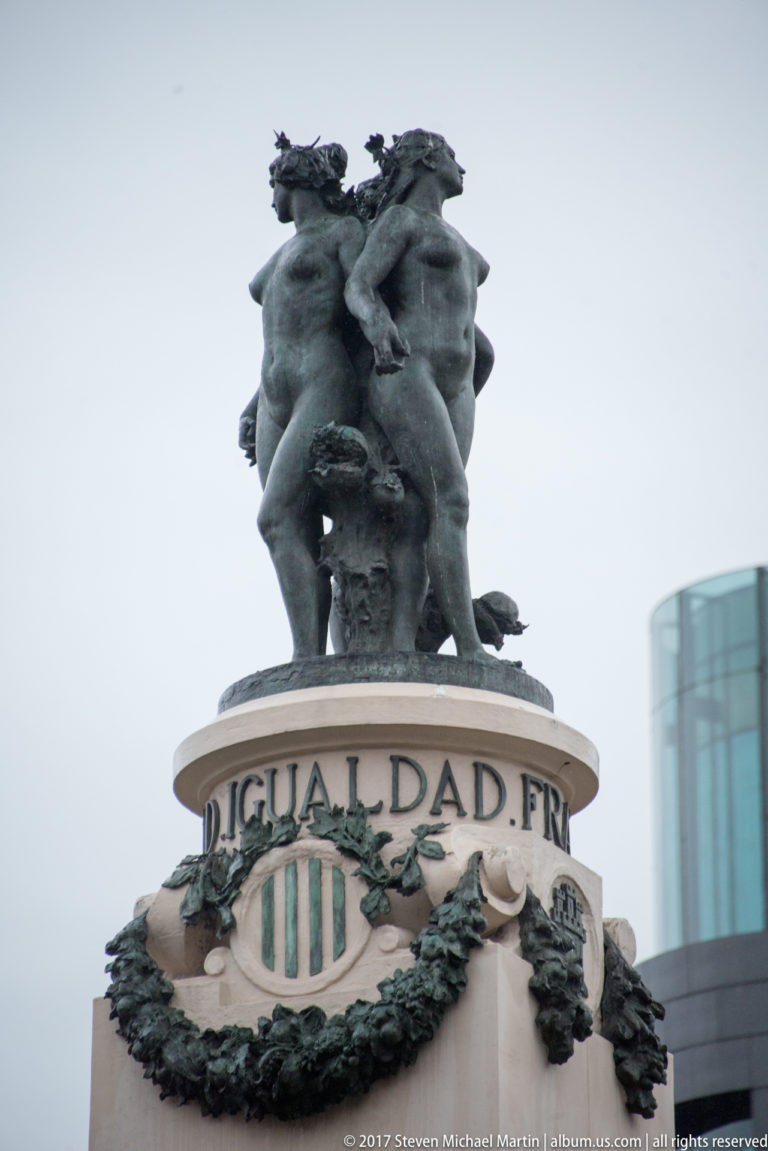 Bronze allegorical figures atop a stone pedestal inscribed “IGUALDAD,” with garlands and a modern building behind.