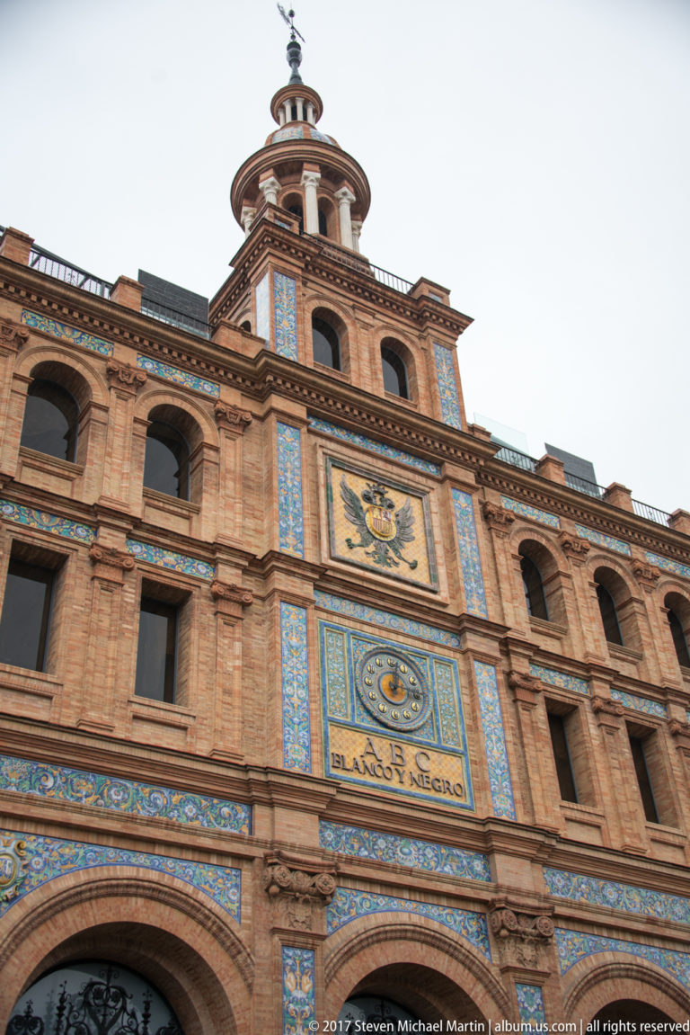 Upward view of Las Ventas bullring with brick arches, blue-and-white tile panels, heraldic decoration, and rooftop pavilion.