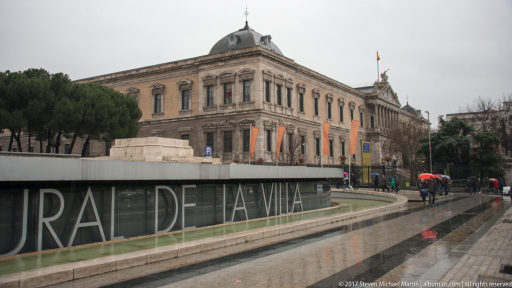 Long side view of the National Library of Spain beside wet pavement, trees, and pedestrians carrying umbrellas.