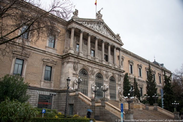 Angled view of the National Library of Spain showing its columned portico, arched entrances, steps, trees, and gardens.