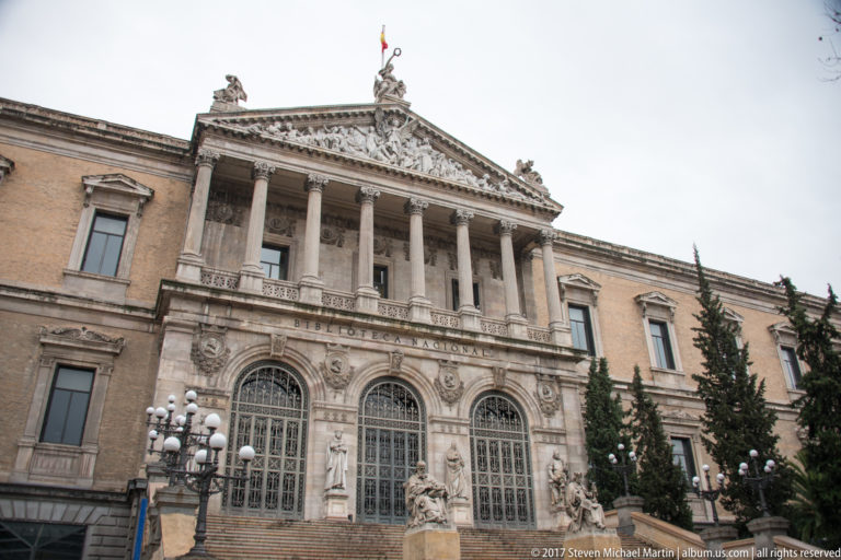 Front façade of Spain’s National Library with Corinthian portico, arched entrances, broad steps, sculptures, and Spanish flag.