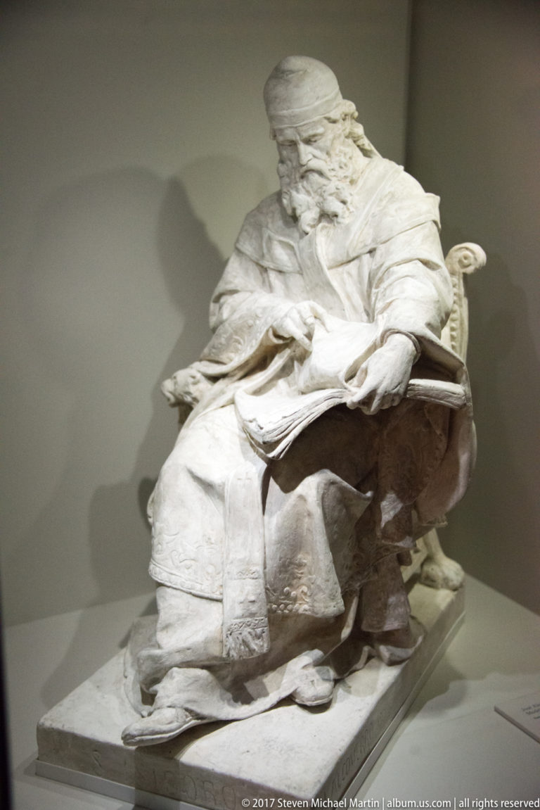 White marble statue of an elderly bearded man seated in robes and looking down at a tablet or book.
