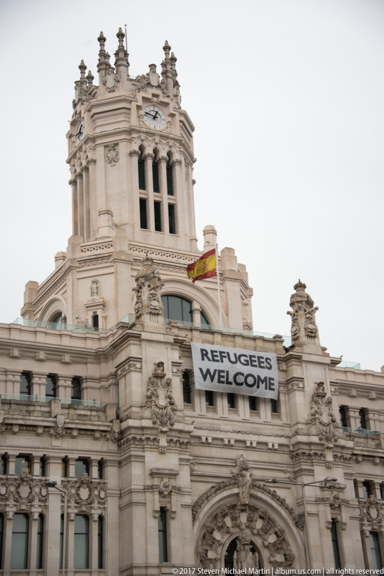 Ornate white façade and tower of Palacio de Cibeles with Spanish flag and a large “REFUGEES WELCOME” banner.