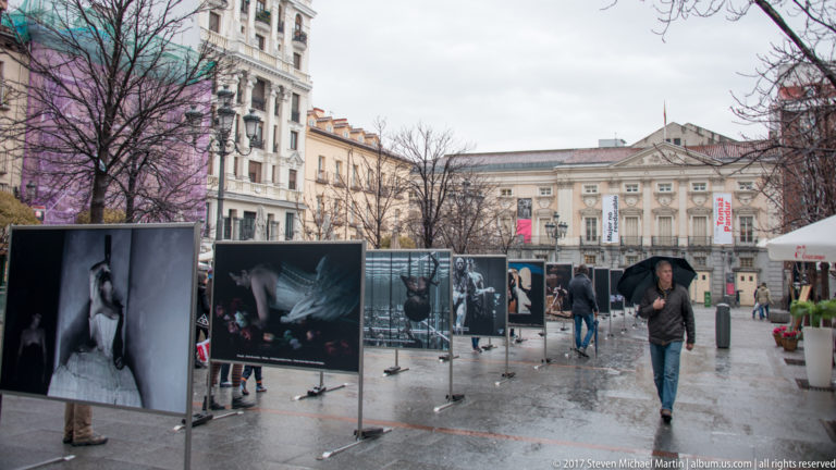 Outdoor photography panels and pedestrians on a wet Madrid square lined by historic buildings and leafless trees.