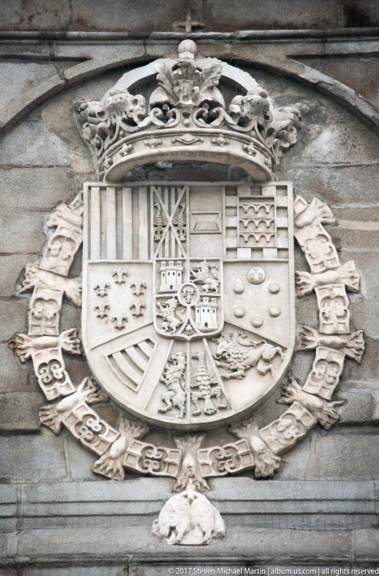 Carved stone coat of arms with royal crown, quartered shield, scrollwork, and surrounding emblems on a Madrid façade.