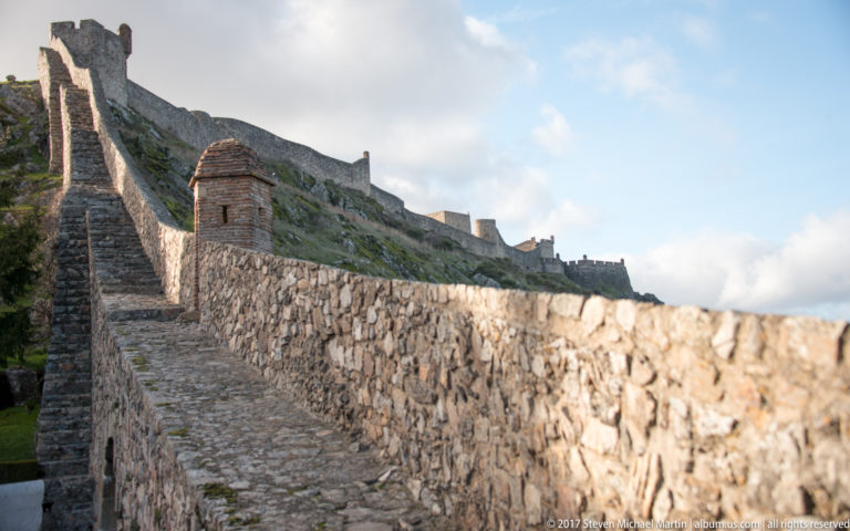 Cobbled rampart walk follows a long stone parapet toward higher fortifications on Marvão’s rocky slope.