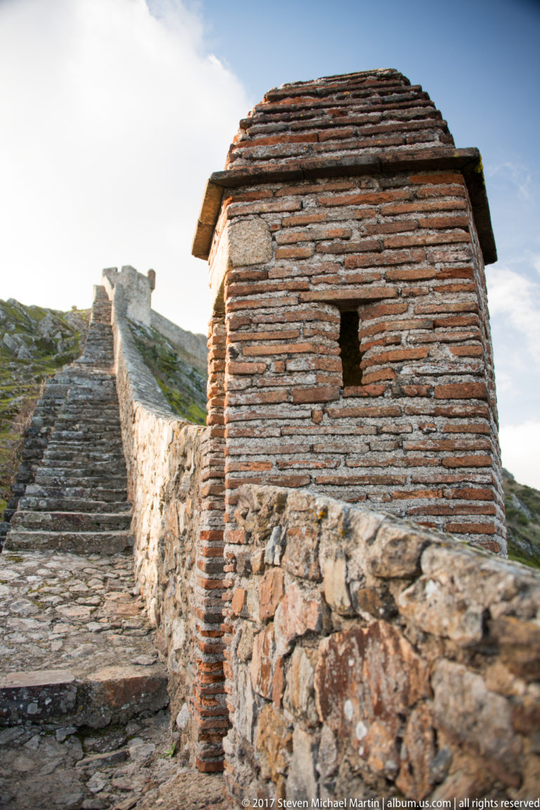 Small brick-and-stone roofed defensive projection with a narrow slit beside stone rampart steps.