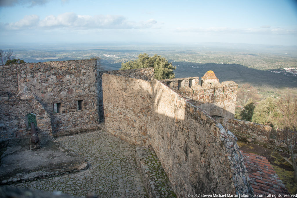 Cobbled castle courtyard lies behind a long stone wall with the distant plain visible beyond.