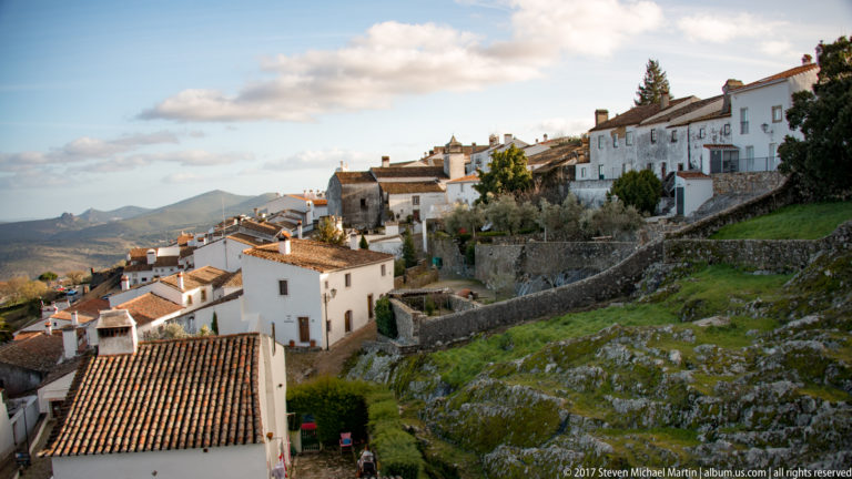 Sunlit white houses step down Marvão’s rocky hillside behind stone walls, gardens, and distant mountains.