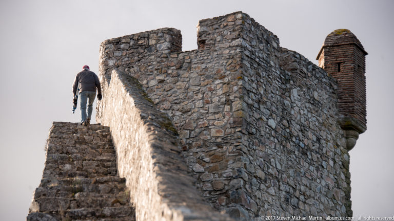 Visitor climbs narrow stone steps along a high masonry wall with a projecting defensive feature at right.