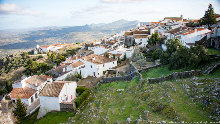 Whitewashed houses and red roofs descend a rocky Marvão hillside between stone terraces with mountains beyond.
