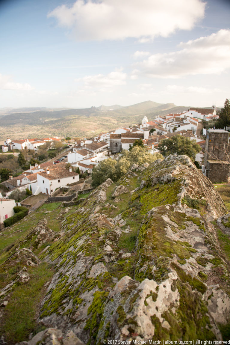 Rocky summit slopes toward scattered white buildings and a broad sunlit plain below Marvão.
