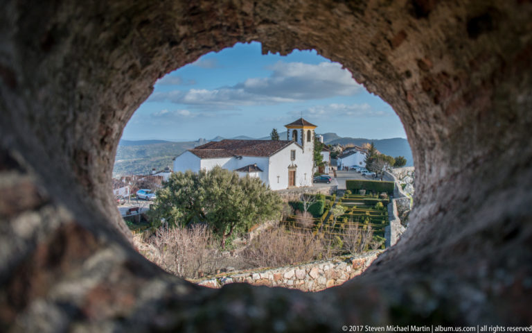 White church and terraced hedges seen through an irregular opening in a thick stone wall.
