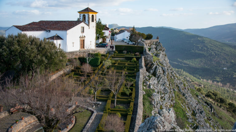 Whitewashed hilltop church overlooks geometric hedged gardens and a steep wooded valley in Marvão.