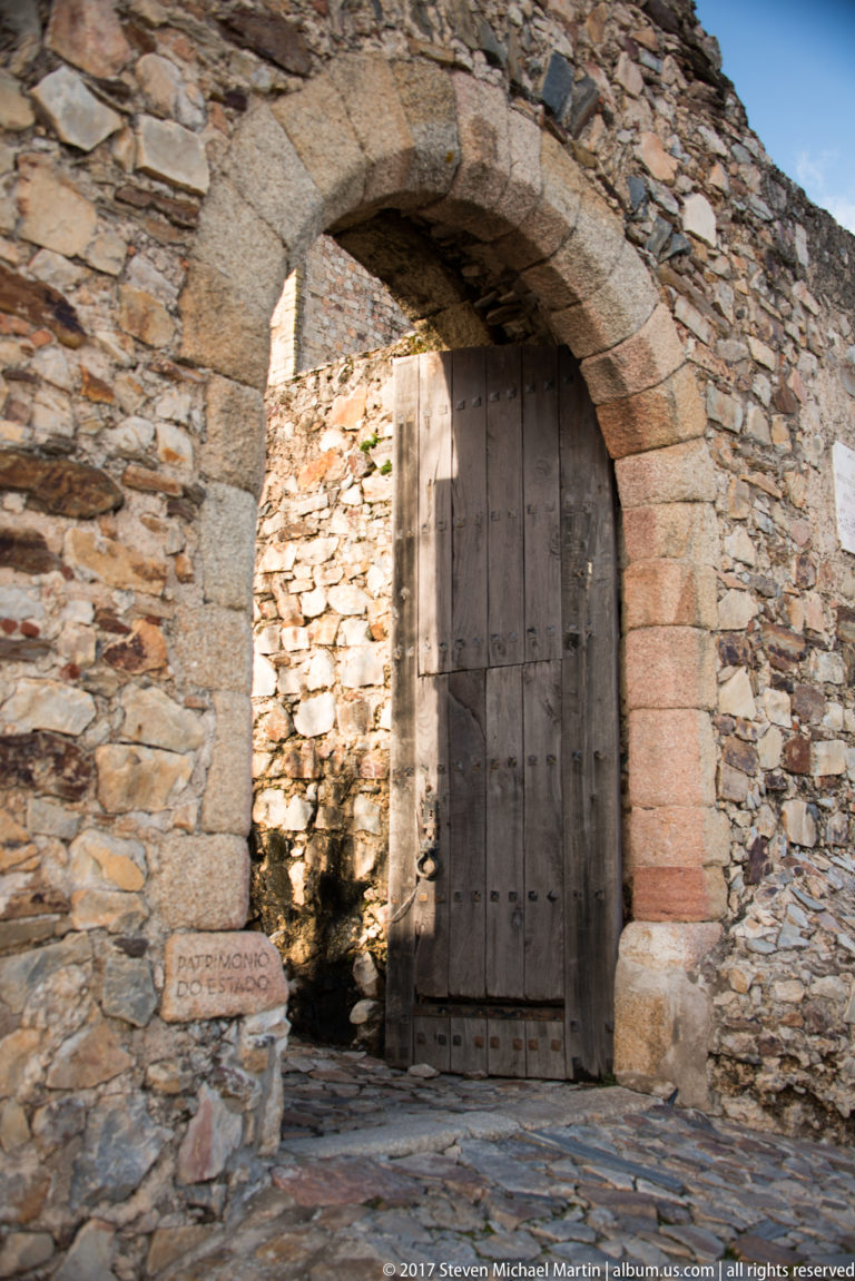 Large timber door stands partly open beneath a dressed-stone arch set in rough rubble masonry.