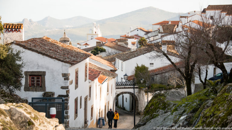 Whitewashed Marvão houses line a steep lane with two pedestrians, tiled roofs, church towers, and mountains beyond.