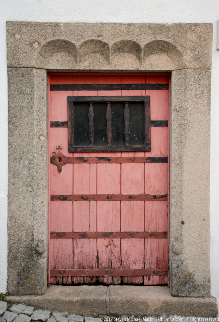 Weathered red plank door with a narrow barred window set in a thick gray granite surround.