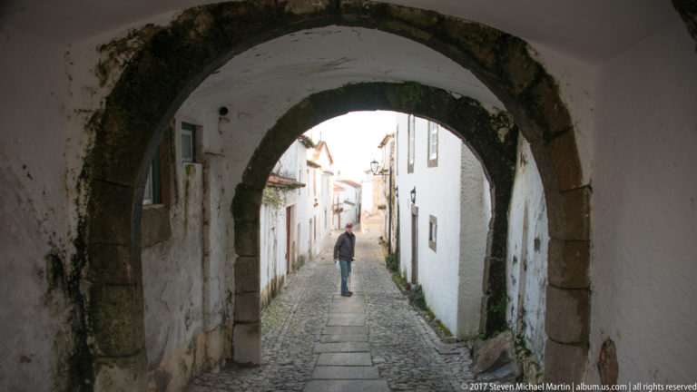 Dark stone arch framing a narrow whitewashed lane with a lone pedestrian walking toward sunlight.