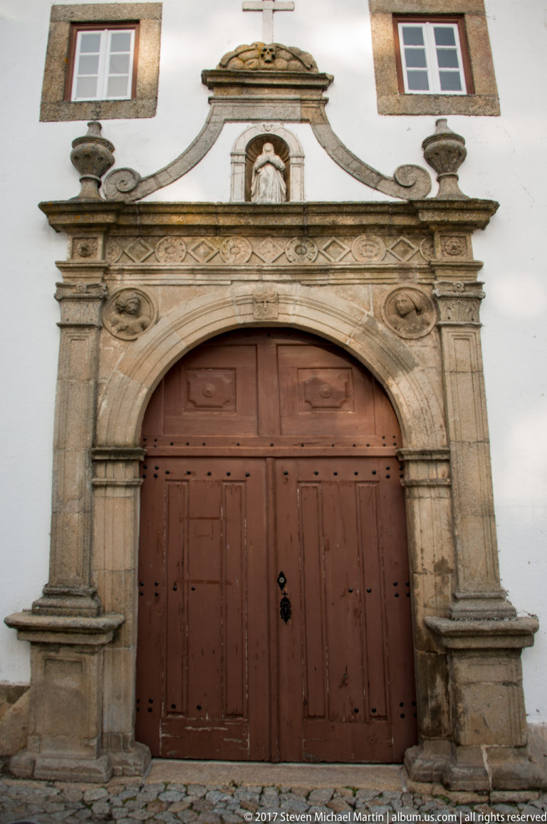 Large wooden double doors within an ornate carved-stone round-arched church portal topped by a curved pediment and cross.
