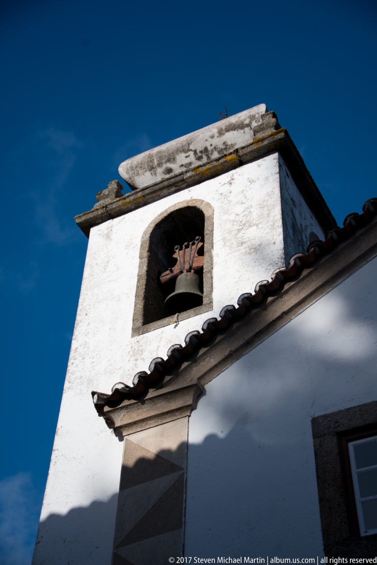 Low-angle view of a single bell hanging in an arched opening of a whitewashed church belfry beneath blue sky.