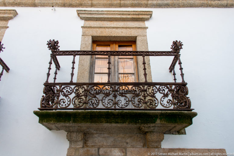 Ornate wrought-iron balcony with scrolling patterns in front of a tall granite-framed window on a white wall.