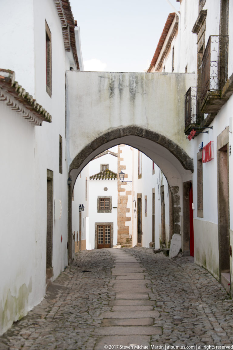 Broad stone arch spanning a cobbled lane between whitewashed houses with balconies and granite trim.