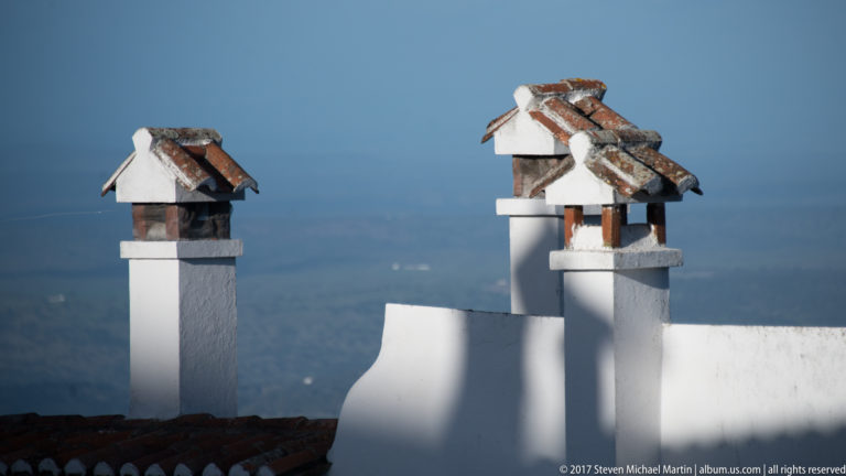 Three whitewashed chimney stacks topped with small pitched red-tile caps against a distant blue landscape.