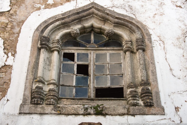 Weathered carved stone window with paired colonnettes, decorative capitals, lobed arch, and broken multi-pane glazing in a white wall.