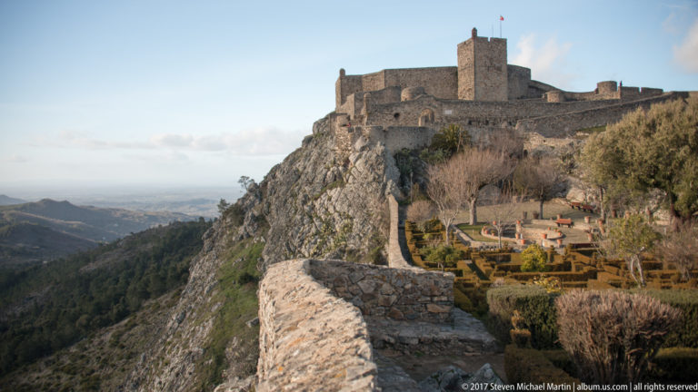 Curving stone wall leading toward Marvão Castle above a garden terrace and steep mountain slope.