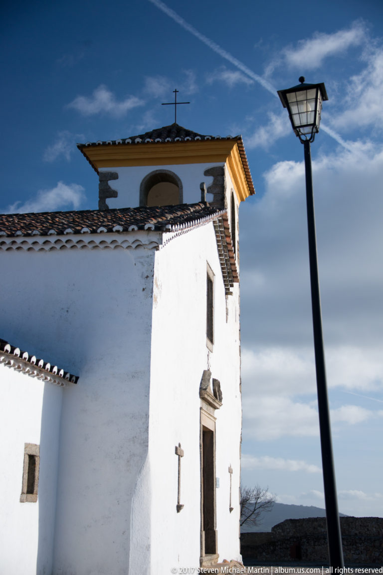 Whitewashed church wall with stone-framed doorway and upper arched belfry opening beside a modern street lamp.
