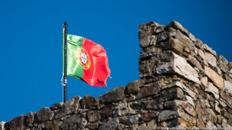 Portuguese green-and-red flag flying above a rough stone battlement against a deep blue sky.