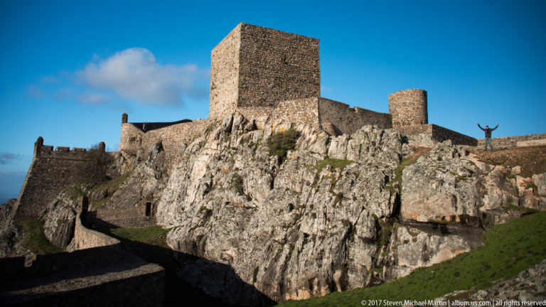 Square castle keep and round tower above a steep granite outcrop, with a small visitor raising both arms on the ridge.