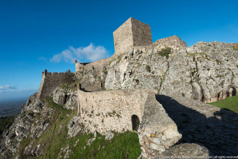 Square stone keep on jagged granite above layered defensive walls, a round tower, and a small arched opening.