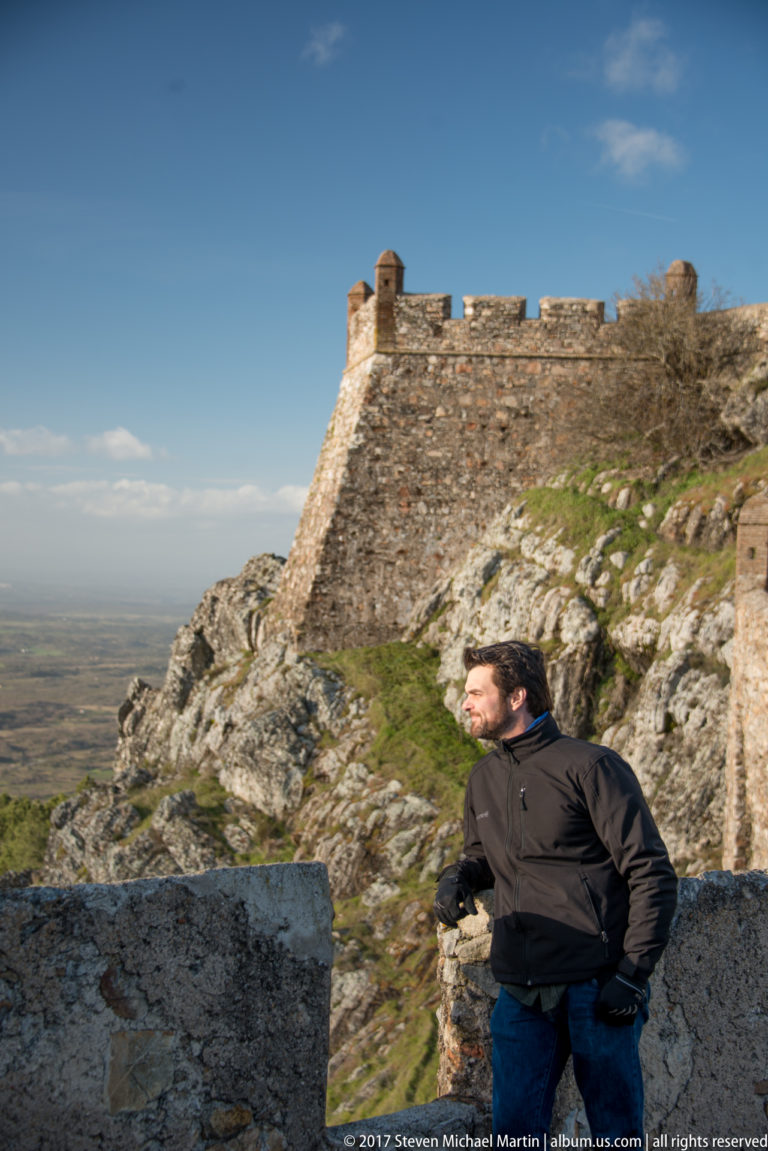 Visitor standing on granite below a massive sloping castle wall and parapet, with distant plain at left.