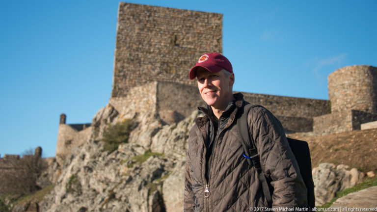 Smiling visitor in a dark jacket and red cap standing before the square keep and stone walls of Marvão Castle.