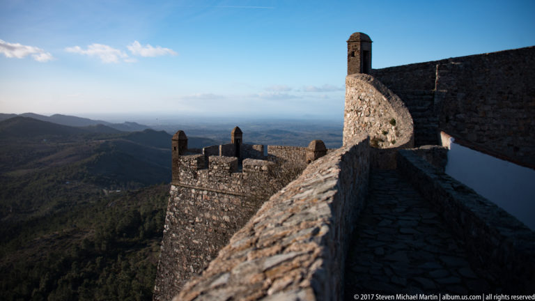 Narrow stone rampart walk beside crenellated walls and small roofed turrets, overlooking dark mountain ridges.